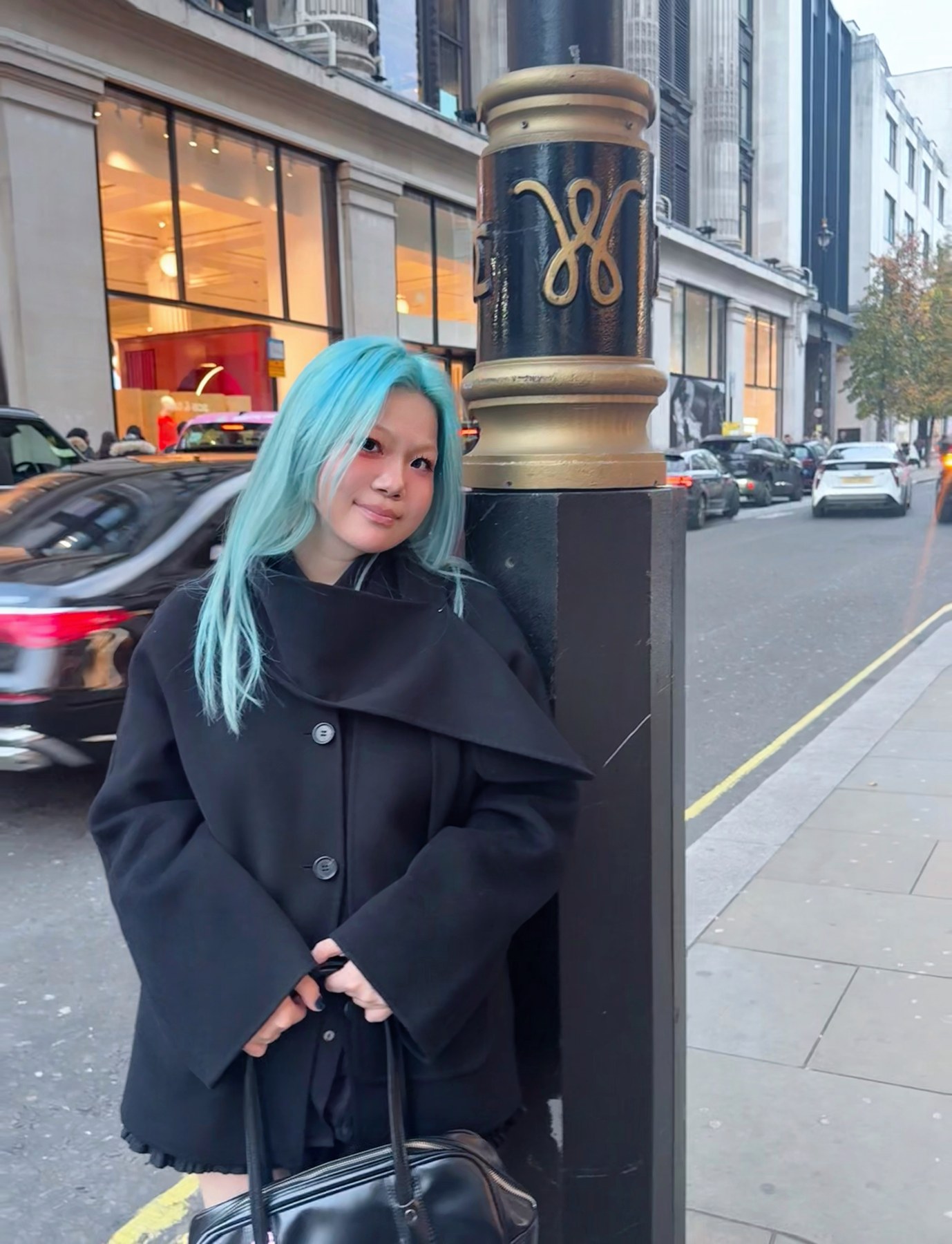 Elaine with blue hair, wearing a black coat on a city street